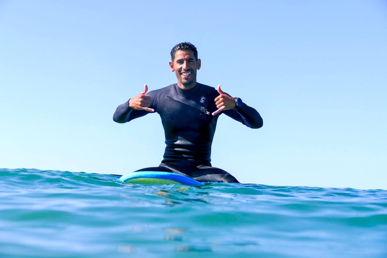 Smiling surfer in a black wetsuit sitting on a surfboard in calm blue ocean