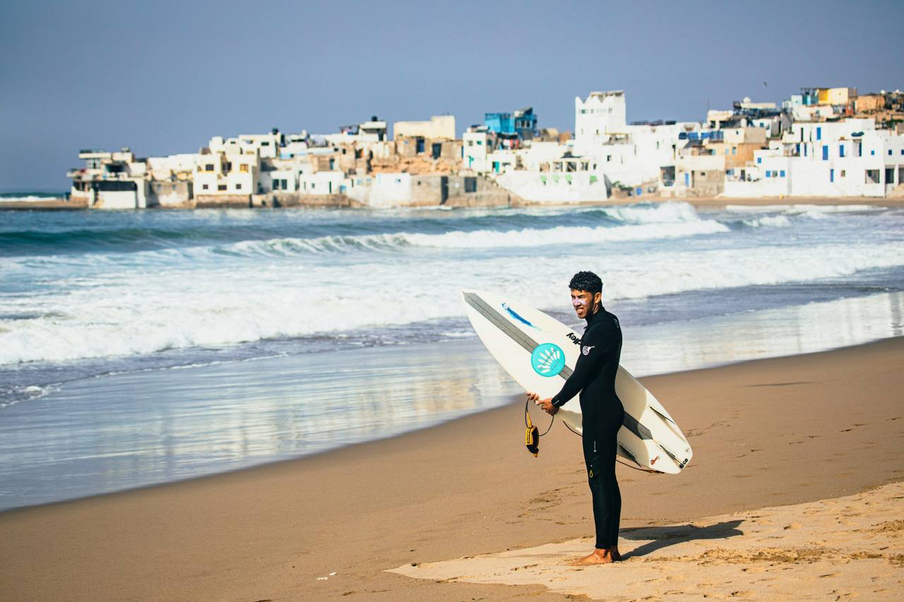 Surfer holding a board on the beach with the coastal town behind them
